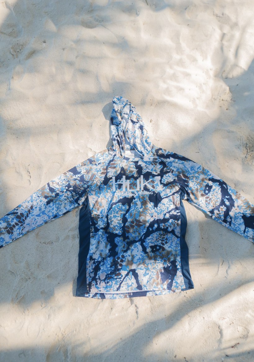 A HUK sun hoodie laid out on white sand in dappled shade